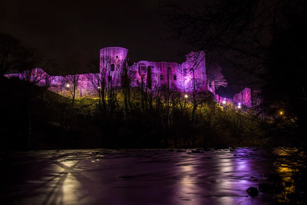 A castle illuminated by purple lights at night, surrounded by trees and reflected in a calm body of water below. - Home Instead