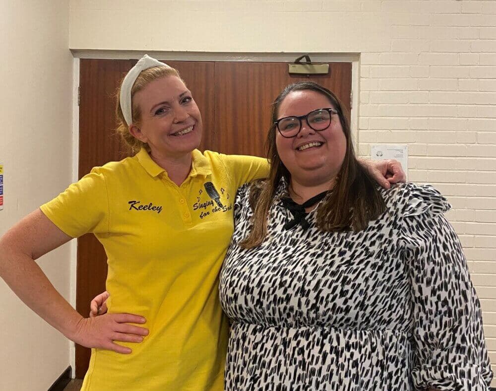 Two women standing together, smiling, one in a yellow polo shirt with "Keeley" on it, the other in a black-and-white patterned dress. - Home Instead