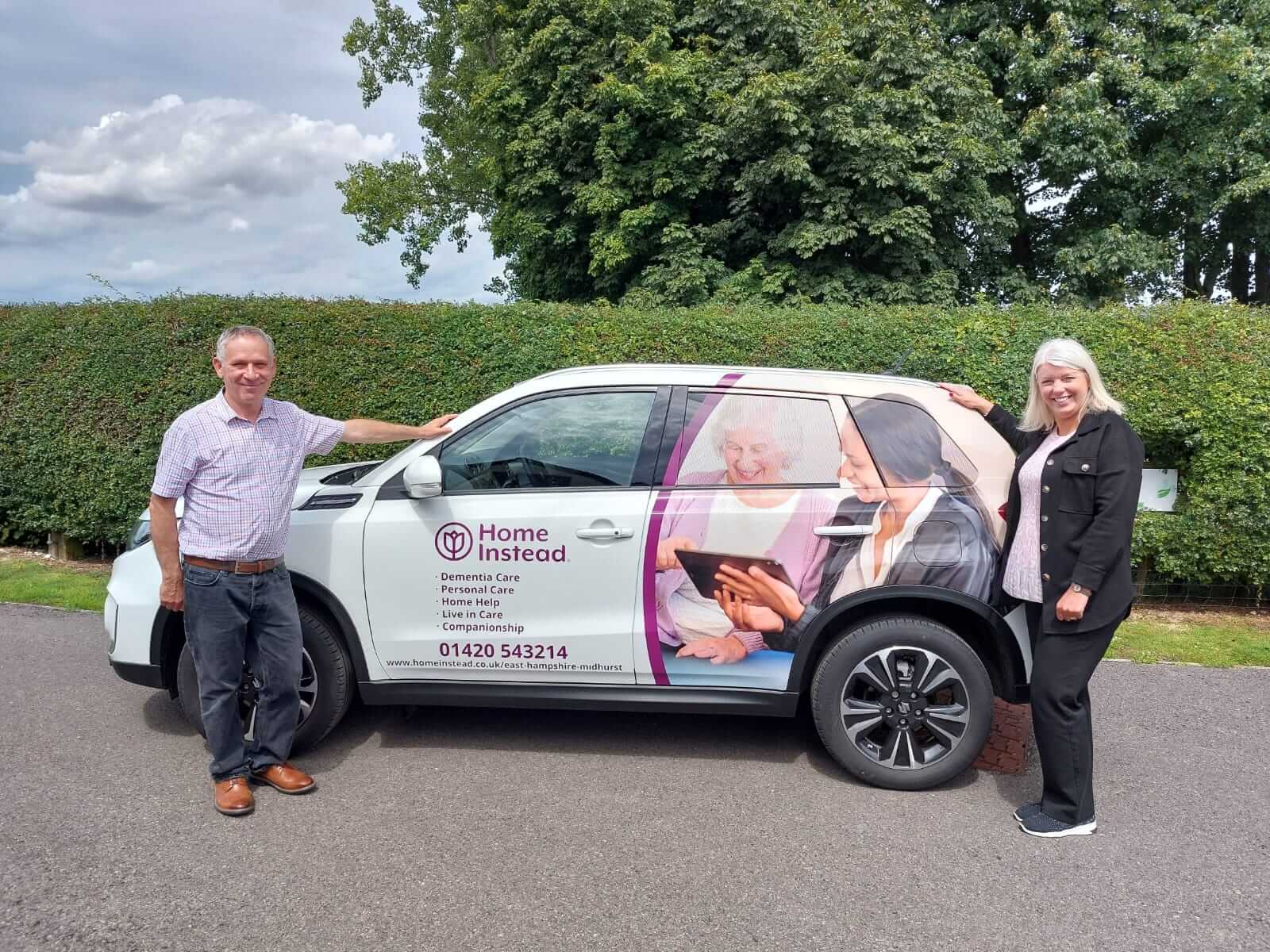 Two people standing next to a car with a Home Instead advertisement, featuring contact info and senior care services. - Home Instead