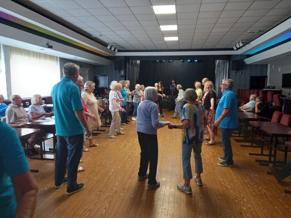 A group of people dancing in a brightly lit hall with tables and chairs arranged on the sides. - Home Instead