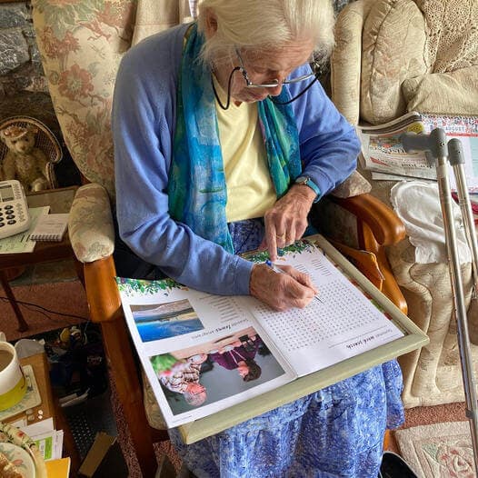 An elderly woman with glasses is seated, solving a word search puzzle book on a tray in a cozy living room. - Home Instead