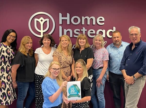 A group of people pose with a "Top 20 Award" sign in front of a Home Instead logo on a purple wall. - Home Instead