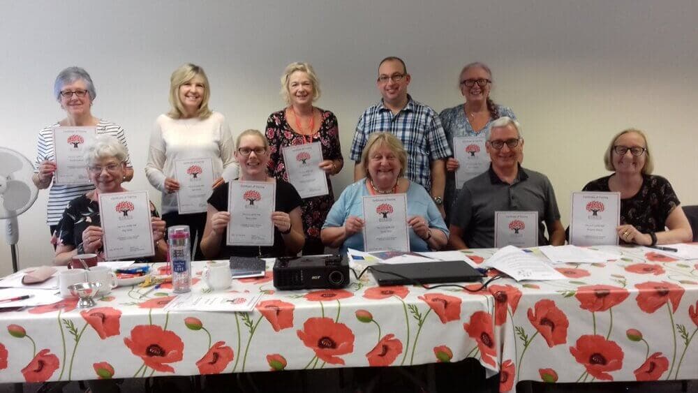A group of people smiling and holding certificates, seated around a table with a poppy-themed tablecloth. - Home Instead