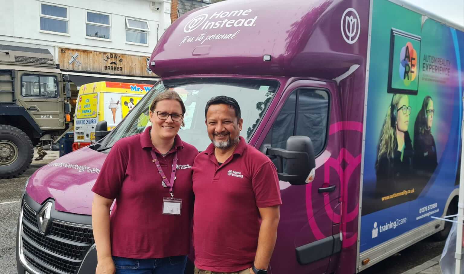 Two smiling people in maroon shirts stand in front of a branded van that says "Home Instead. - Home Instead