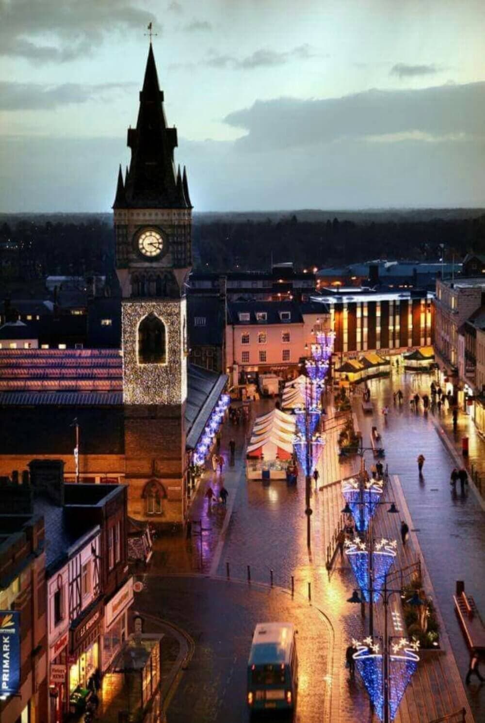 A clock tower and festive lights illuminate a city square with people and a lit Christmas tree at dusk. - Home Instead