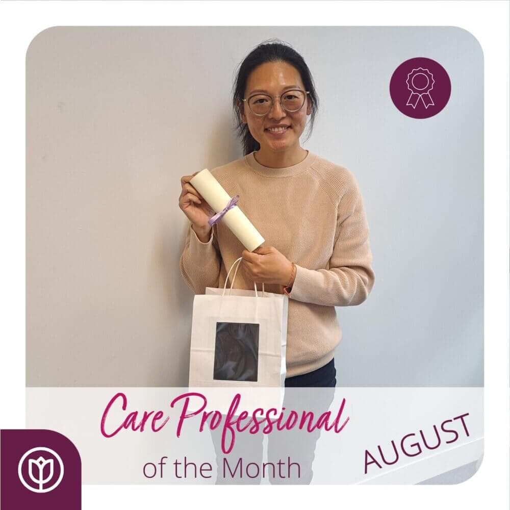 Person holding a certificate and gift bag with "Care Professional of the Month AUGUST" text and award icons. - Home Instead