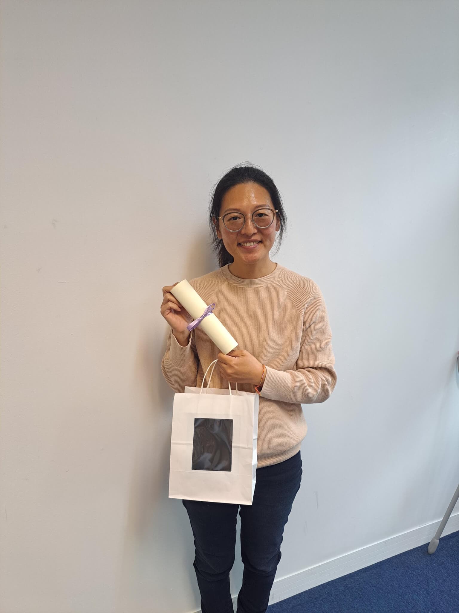 Person smiling, holding a rolled certificate and a gift bag against a white wall background. - Home Instead