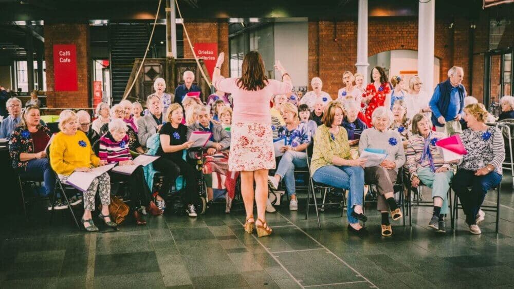 A woman conducts a seated choir of elderly people inside a large building with red banners and brick walls. - Home Instead