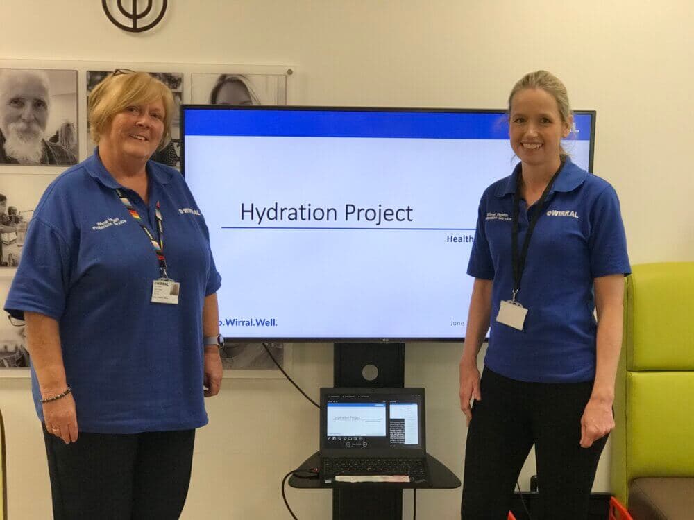 Two women in blue shirts stand beside a screen displaying "Hydration Project" in a room with photos on the wall. - Home Instead