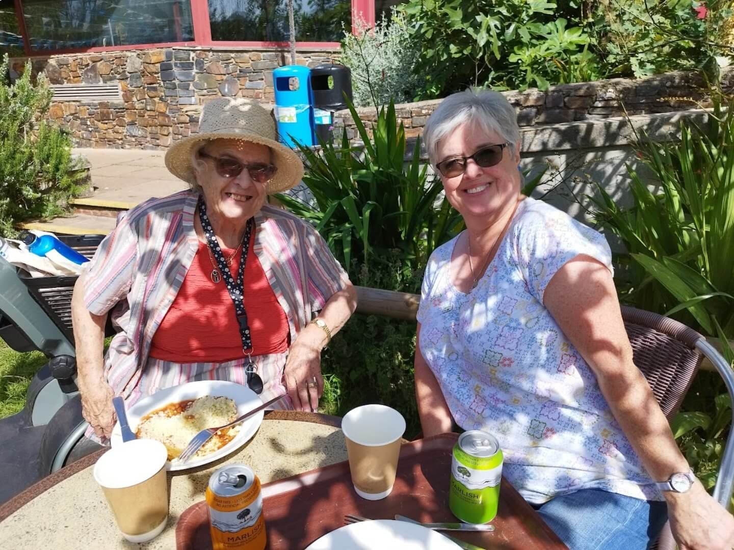 Two women smiling, sitting at an outdoor table with drinks. One wears a sunhat, the other has short gray hair. - Home Instead