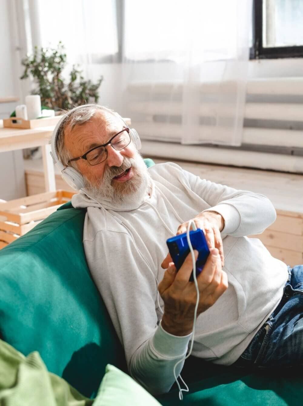 Elderly man with headphones relaxing on a green couch, holding a blue phone, and enjoying music. - Home Instead
