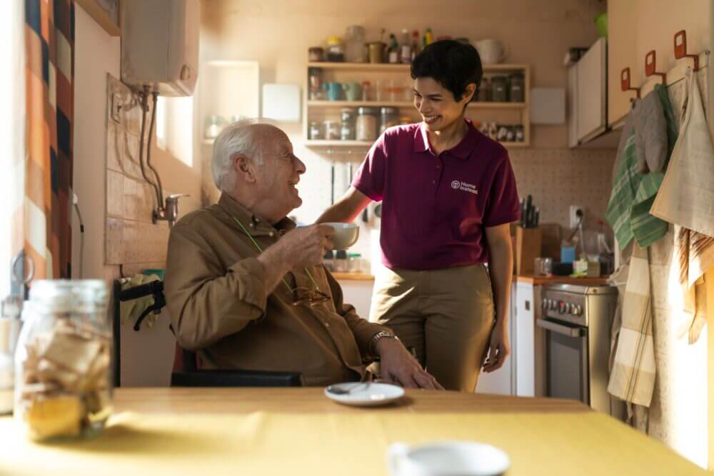 A young woman smiles while standing next to an elderly man sitting at the kitchen table, holding a cup of coffee. - Home Instead