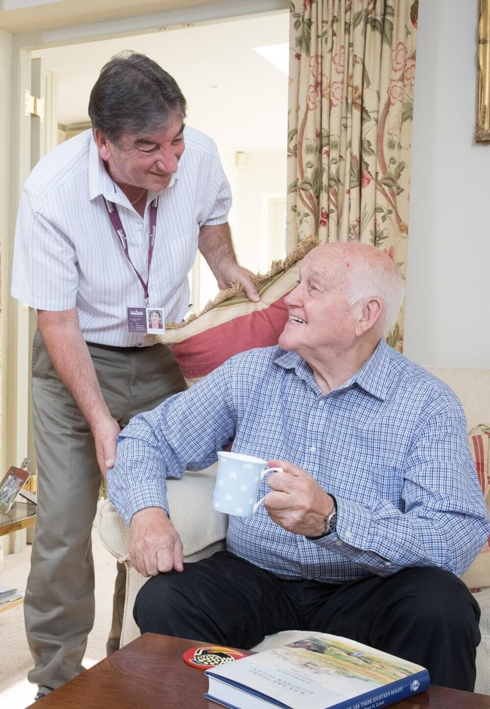 A caregiver converses with an elderly man seated with a mug in hand, both smiling in a brightly lit room. - Home Instead