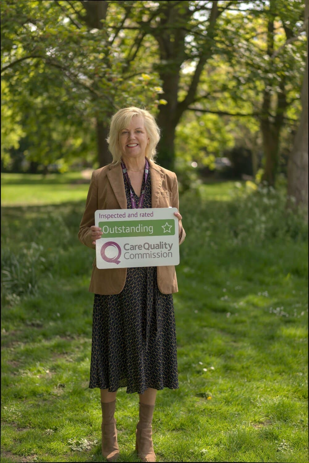 A woman stands outside holding a sign reading "Outstanding Care Quality Commission," smiling with trees in the background. - Home Instead