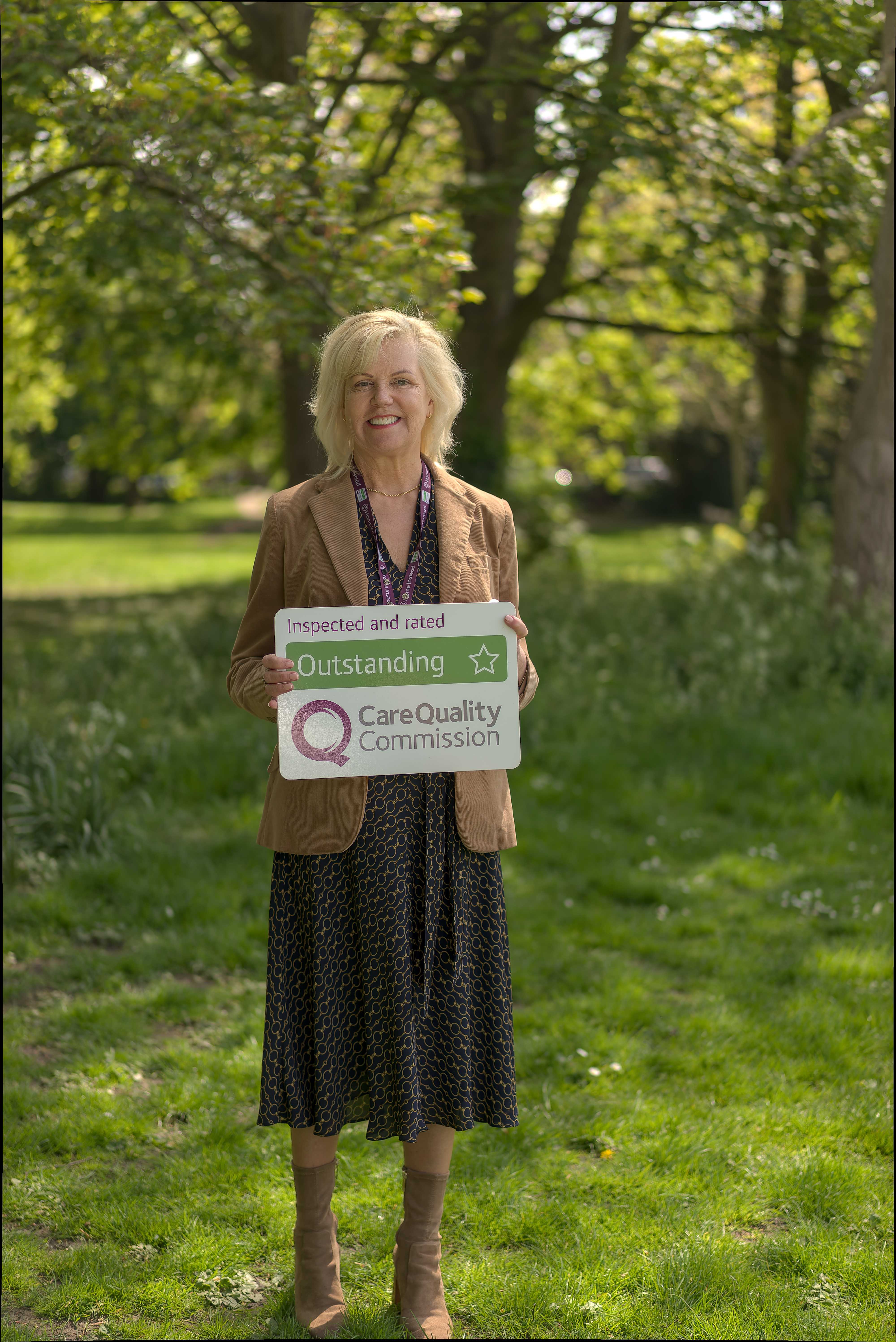 A woman stands outside holding a sign reading "Outstanding Care Quality Commission," smiling with trees in the background. - Home Instead
