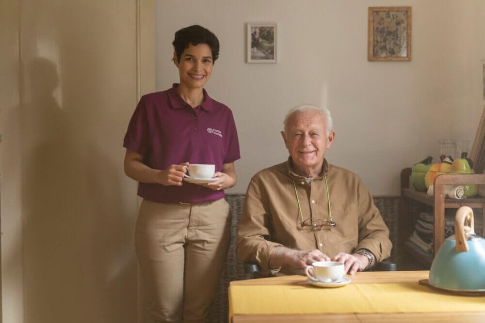 A woman and an elderly man with teacups smile at a table in a warmly lit room with framed pictures on the wall. - Home Instead