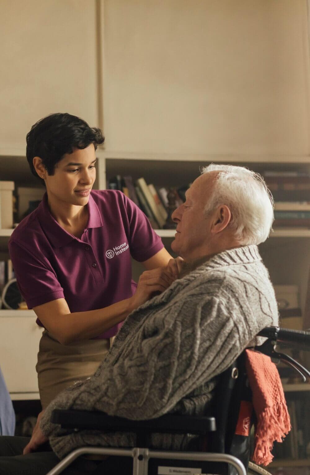 A Care Professional in a purple polo shirt assists an elderly man in a wheelchair with adjusting his knit sweater. - Home Instead Bournemouth & Christchurch