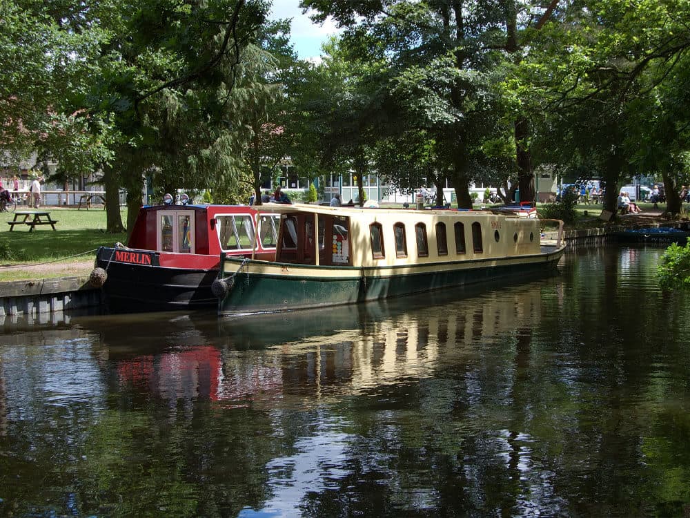 Two canal boats, one red and one beige, moored on a calm river lined with trees on a sunny day. - Home Instead