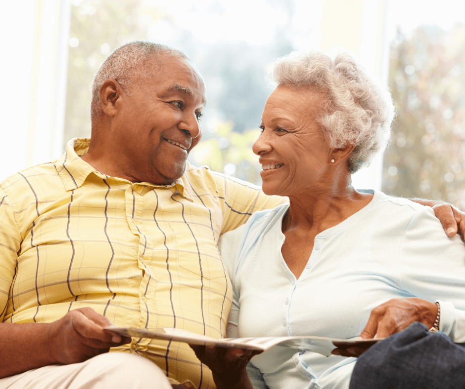 An elderly couple sitting together on a couch, smiling at each other while holding and looking at photographs. - Home Instead