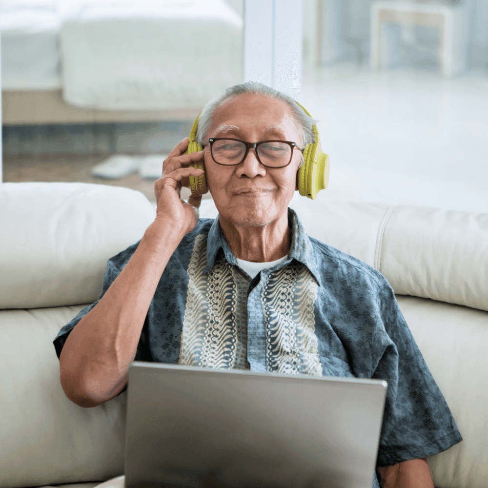 Elderly man wearing headphones and glasses, smiling while sitting on a couch, with a laptop on his lap. - Home Instead