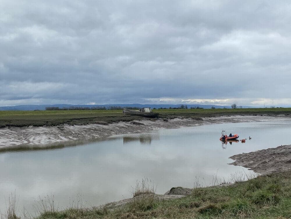 A serene river scene with a small red boat and an old wrecked ship on the grassy shore under a cloudy sky. - Home Instead
