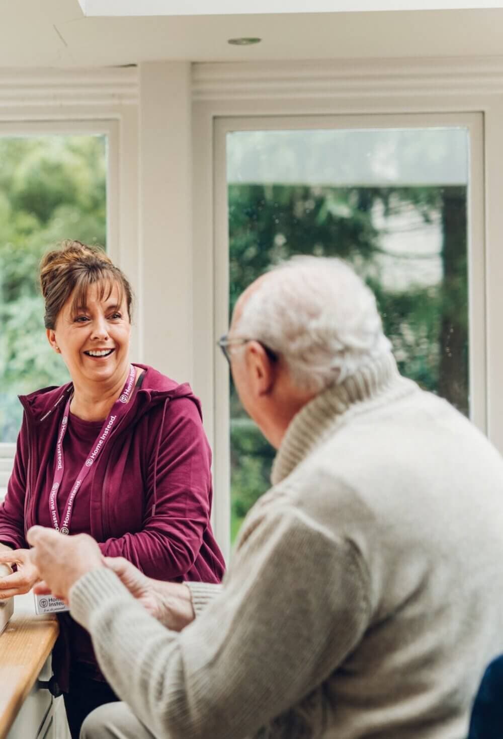 A caregiver in a purple jacket smiling and talking to an elderly man wearing glasses and a beige sweater by a window. - Home Instead