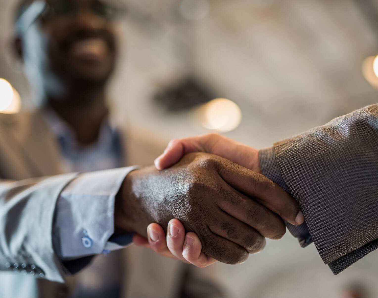 Two people shaking hands, one wearing a light-colored suit and the other in a dark-colored suit, with a blurred background. - Home Instead