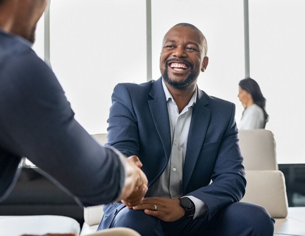 Man in a blue suit smiling and shaking hands with another person in an office setting. - Home Instead
