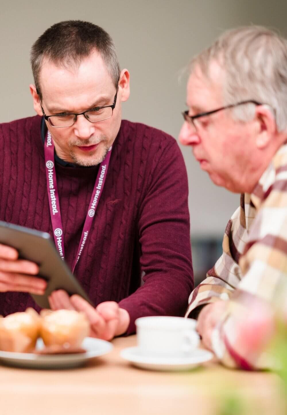 Two men sitting at a table, one showing the other a tablet. A cup and pastries are on the table in front of them. - Home Instead