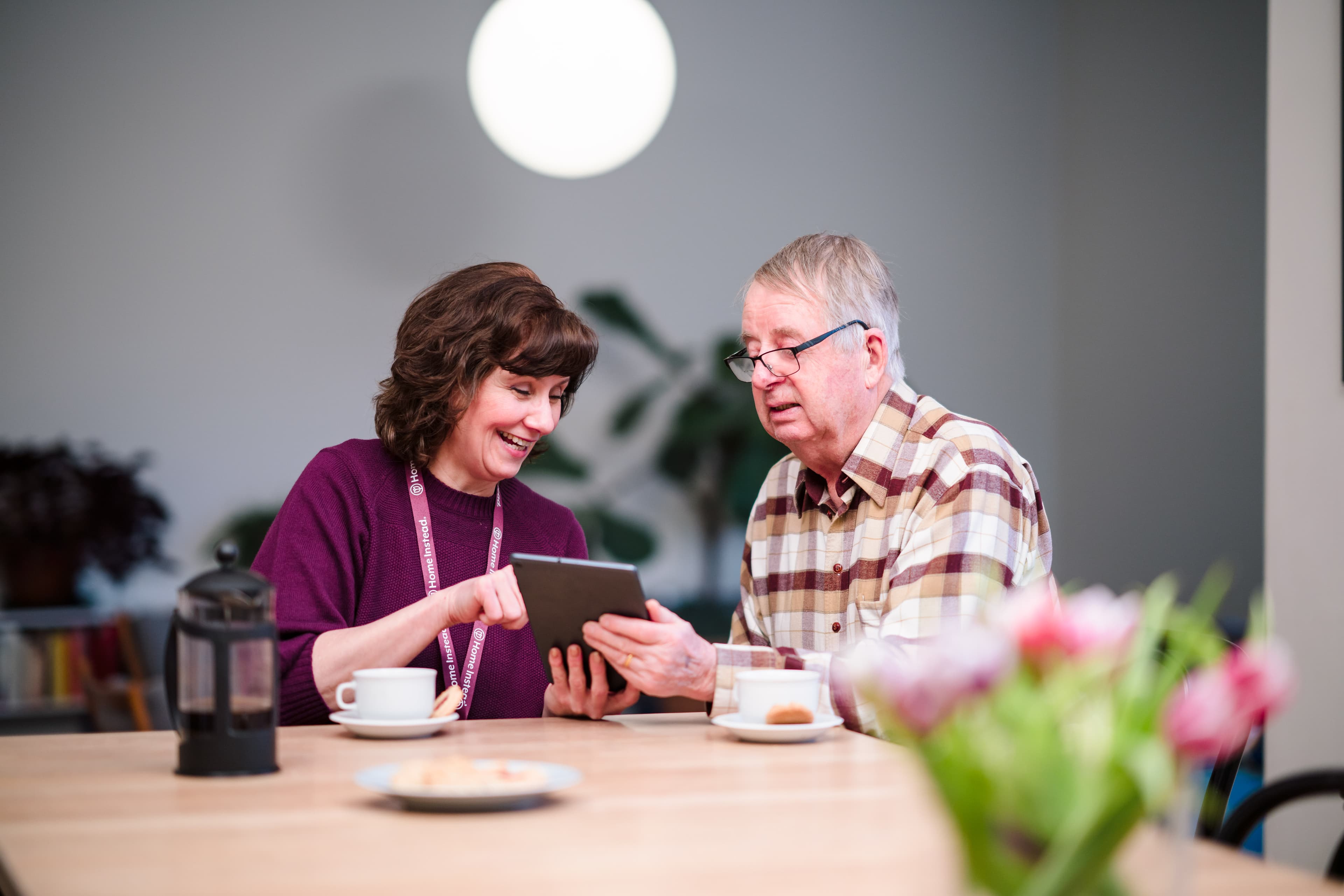 A woman and an elderly man sit at a table, smiling while looking at a tablet together. - Home Instead