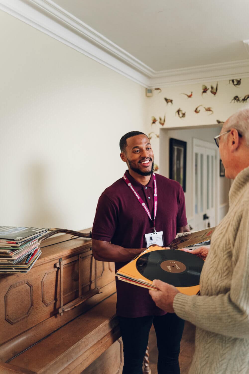 A man in a maroon shirt and ID badge hands a vinyl record to an older man by a wooden piano in a cozy room. - Home Instead