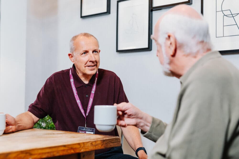 Two elderly men, one wearing an ID badge, sit at a table and chat over coffee in an art-adorned setting. - Home Instead