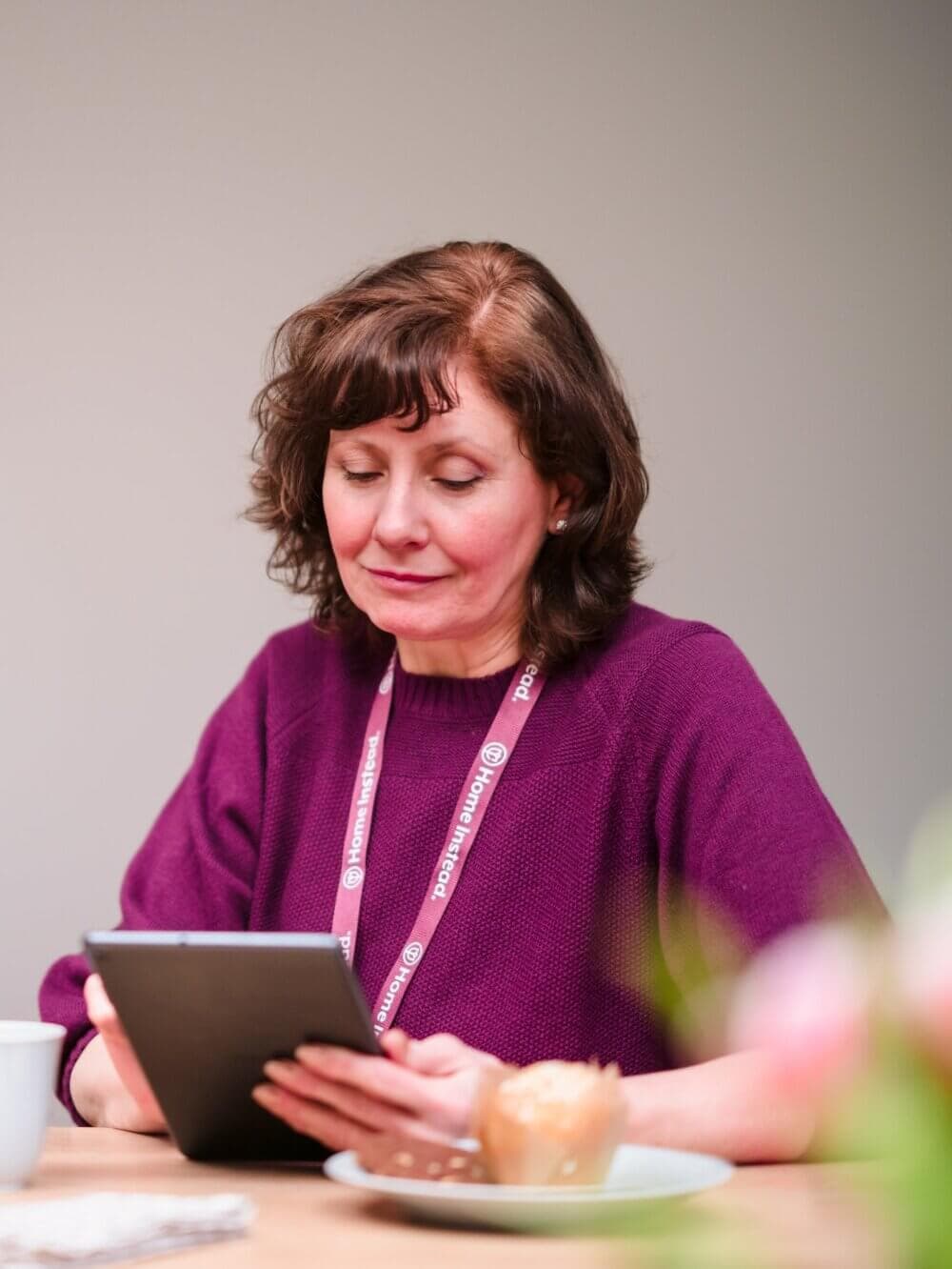 A woman with shoulder-length brown hair reads from a tablet, wearing a purple sweater and a lanyard, sitting at a table. - Home Instead