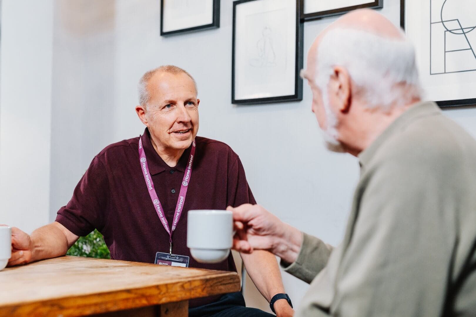 Two older men sit at a table and drink coffee while engaging in a conversation. - Home Instead