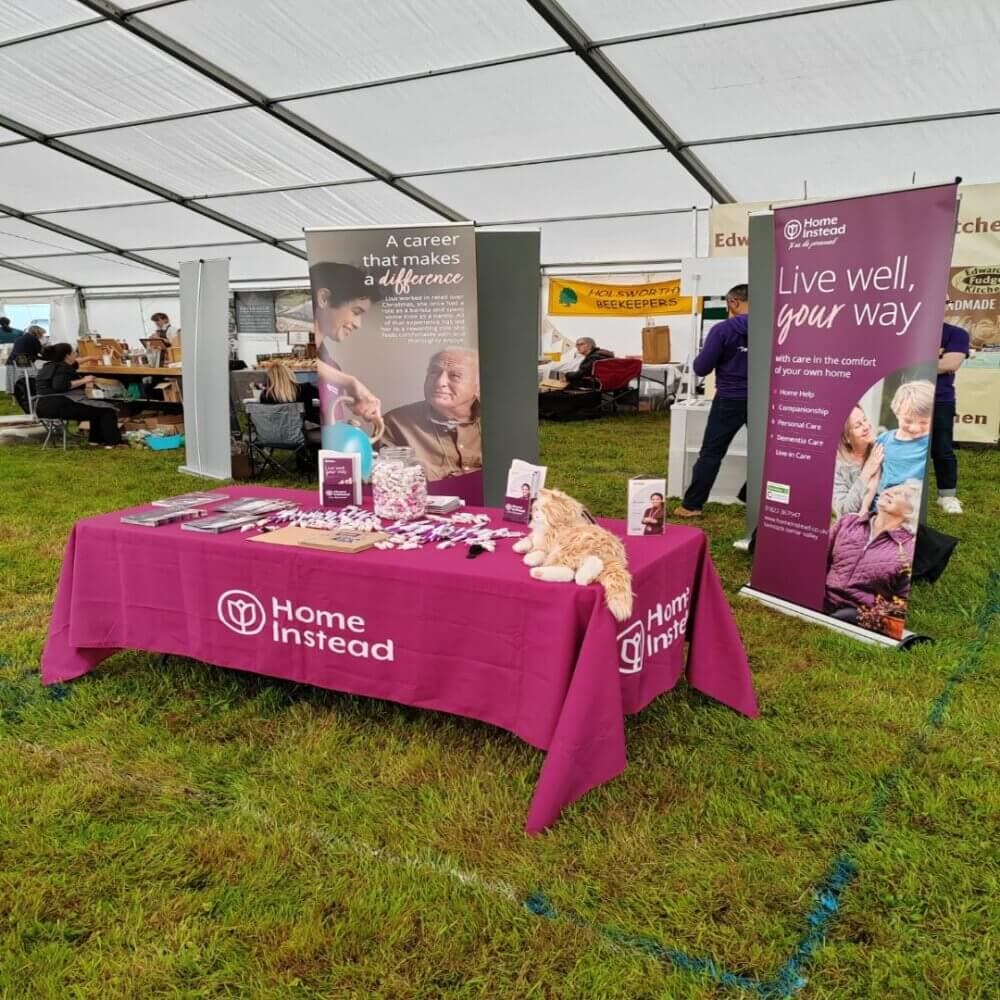 A booth at an outdoor event with banners and a table covered in Home Instead branding, brochures, and promotional items. - Home Instead