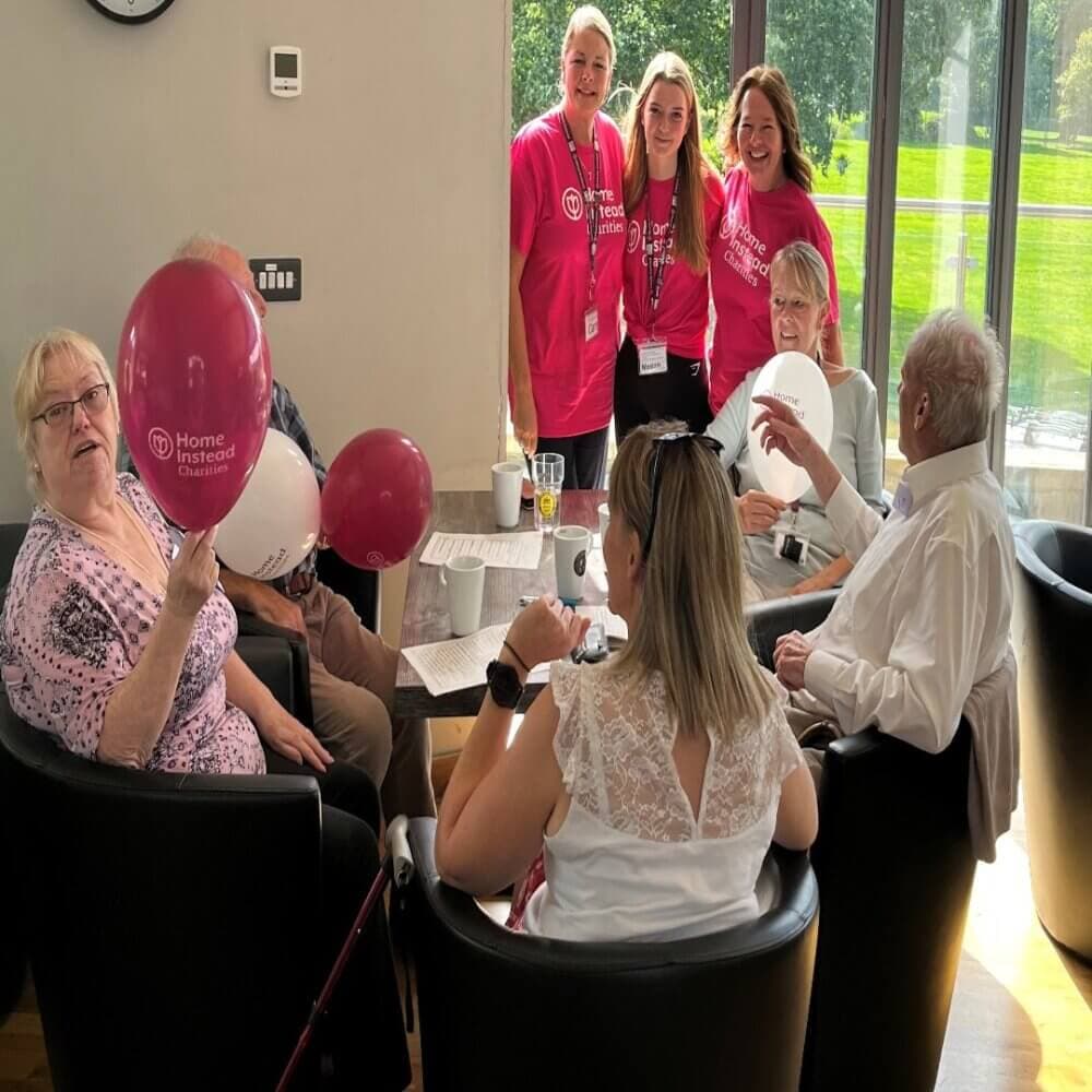 A group of people, some holding balloons, are gathered around a table and smiling for the camera indoors. - Home Instead