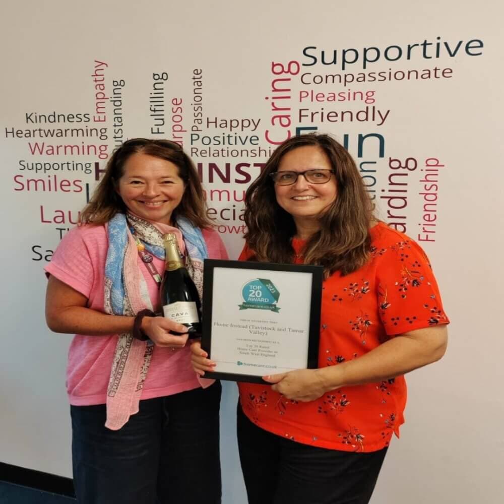 Two women smiling, one holding a bottle of wine, the other holding a framed award, standing against a positive word backdrop. - Home Instead