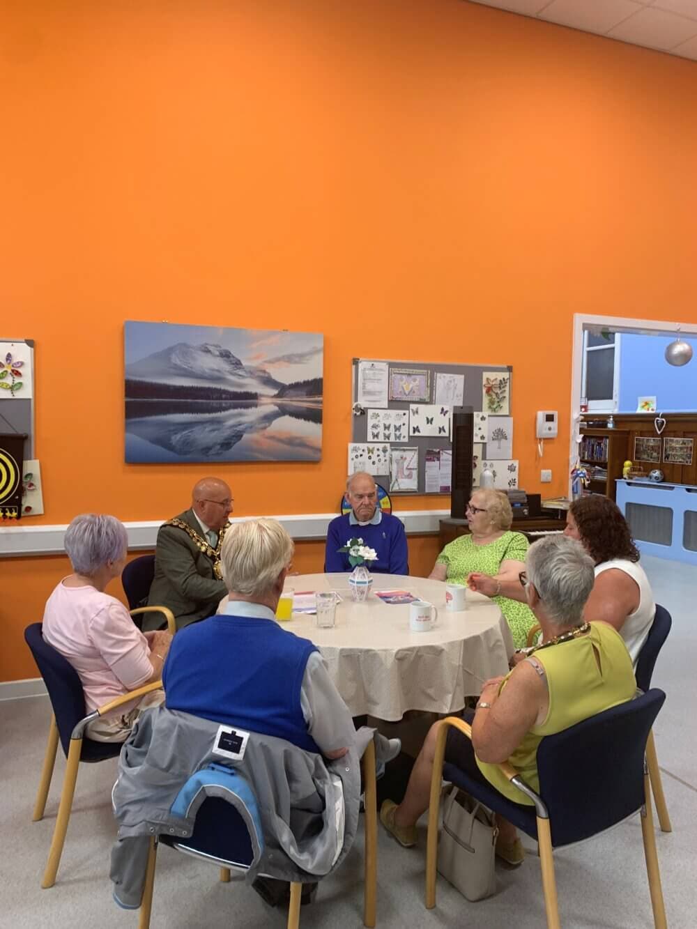 A group of older adults sit around a table in a brightly colored orange room, engaged in lively conversation. - Home Instead