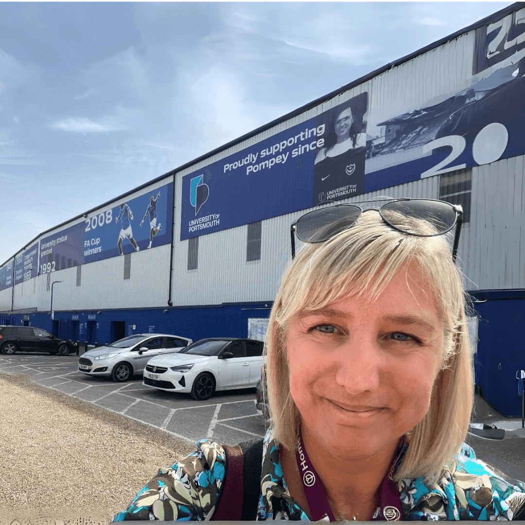 A woman with blonde hair and sunglasses atop her head stands in front of a building with blue and white sports murals. - Home Instead