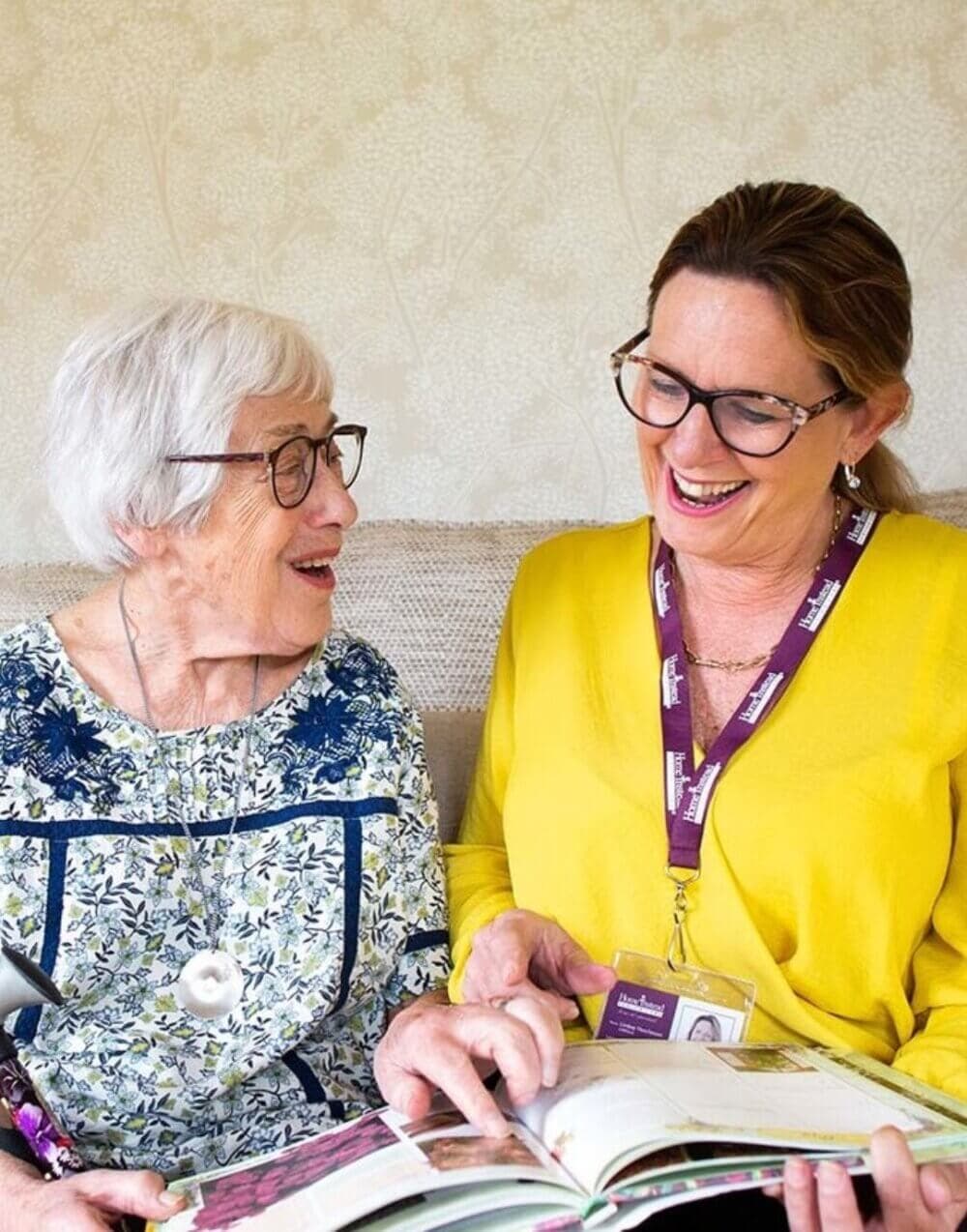 Two women sitting on a couch, happily reading a book together. The older woman is on the left and the younger on the right. - Home Instead