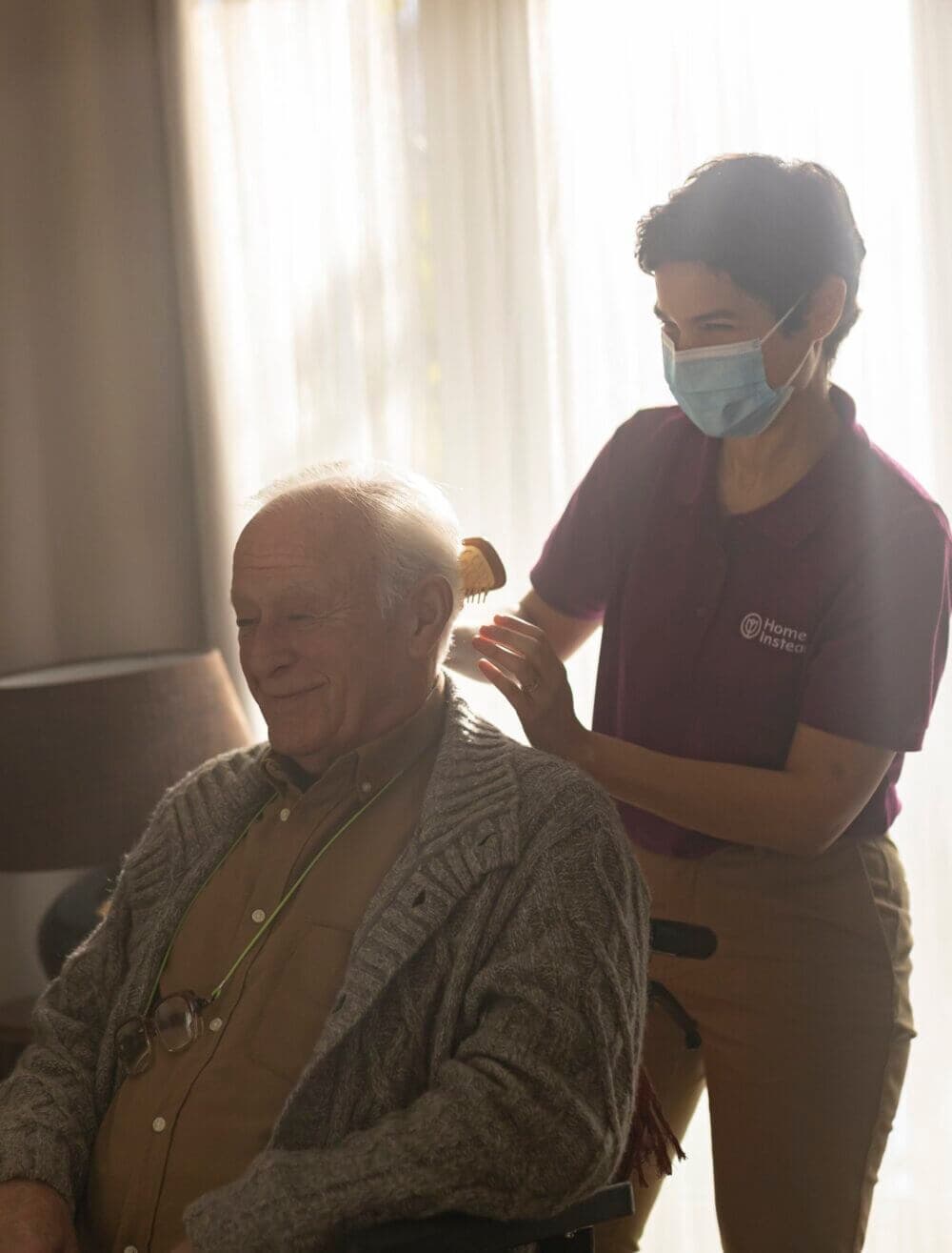 A caregiver wearing a mask brushes the hair of an elderly man seated in a chair in front of a window with curtains. - Home Instead