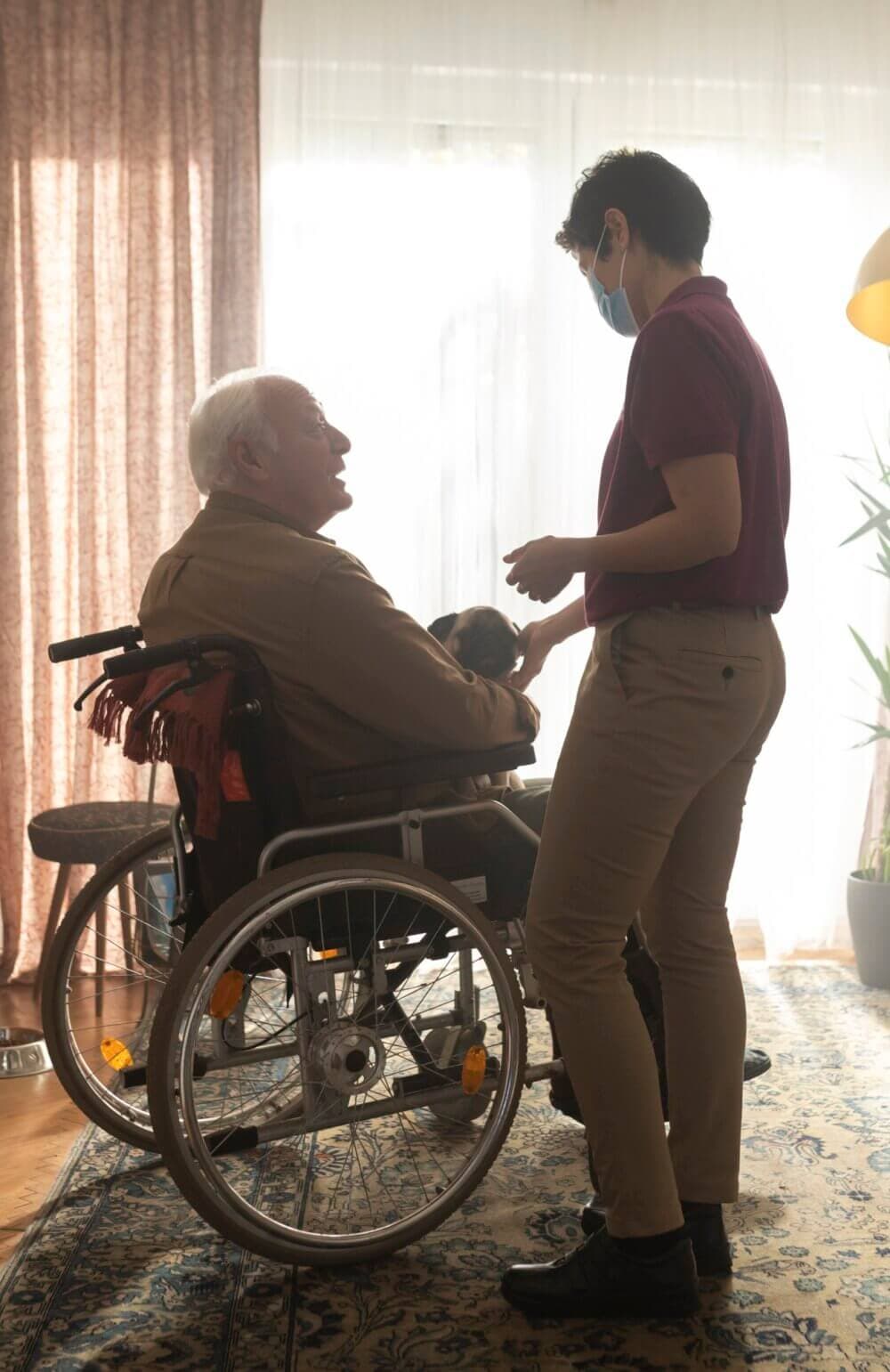 A caregiver in a mask assists a smiling elderly man in a wheelchair in a bright room with curtains. - Home Instead