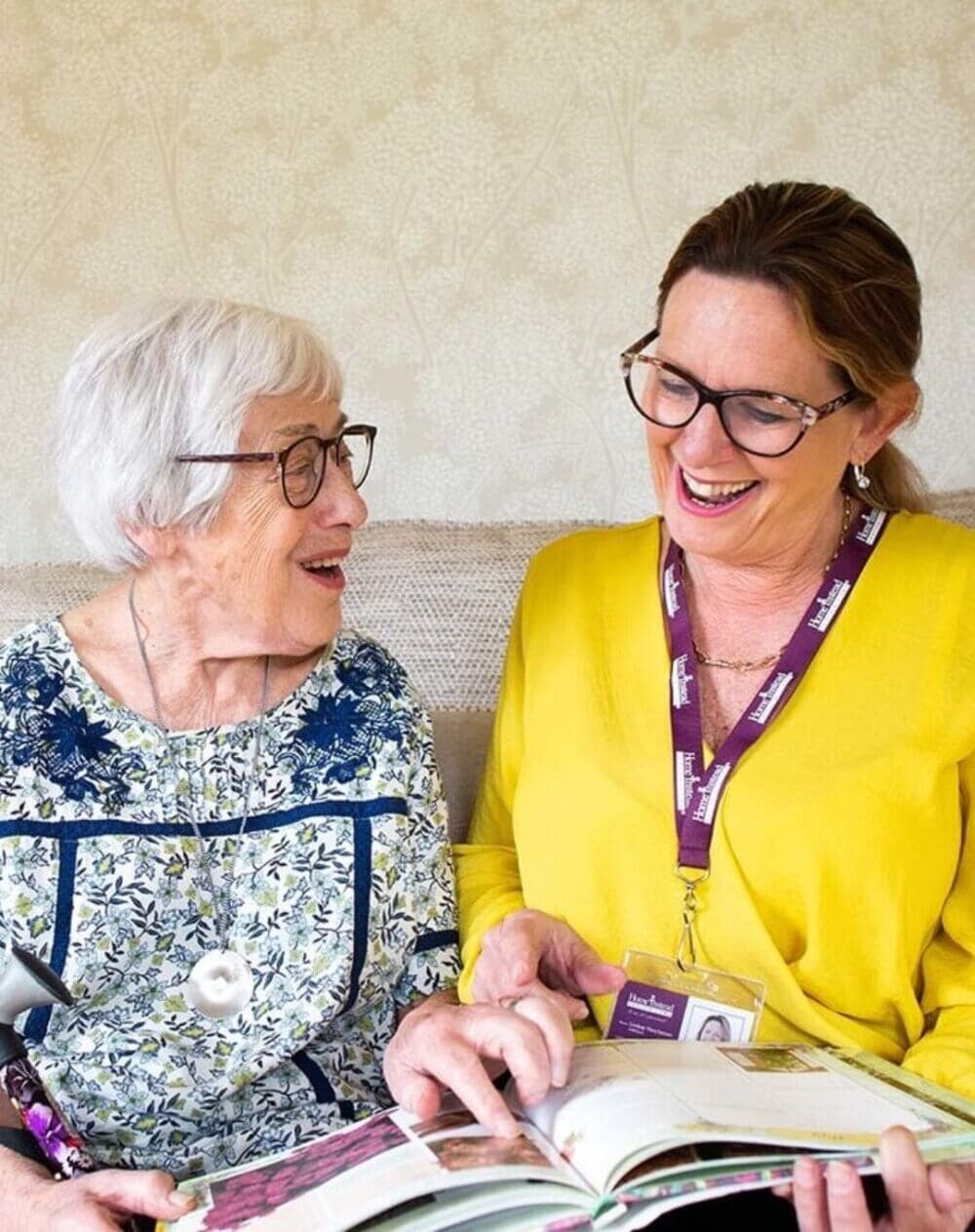 An elderly woman and a caregiver in glasses share smiles while looking at a book together. - Home Instead