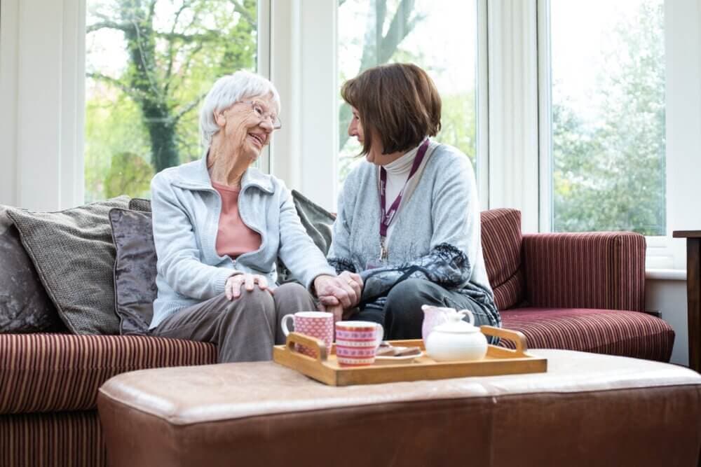Two women sitting on a sofa, smiling at each other with a tray of tea and cups on an ottoman in front of them. - Home Instead