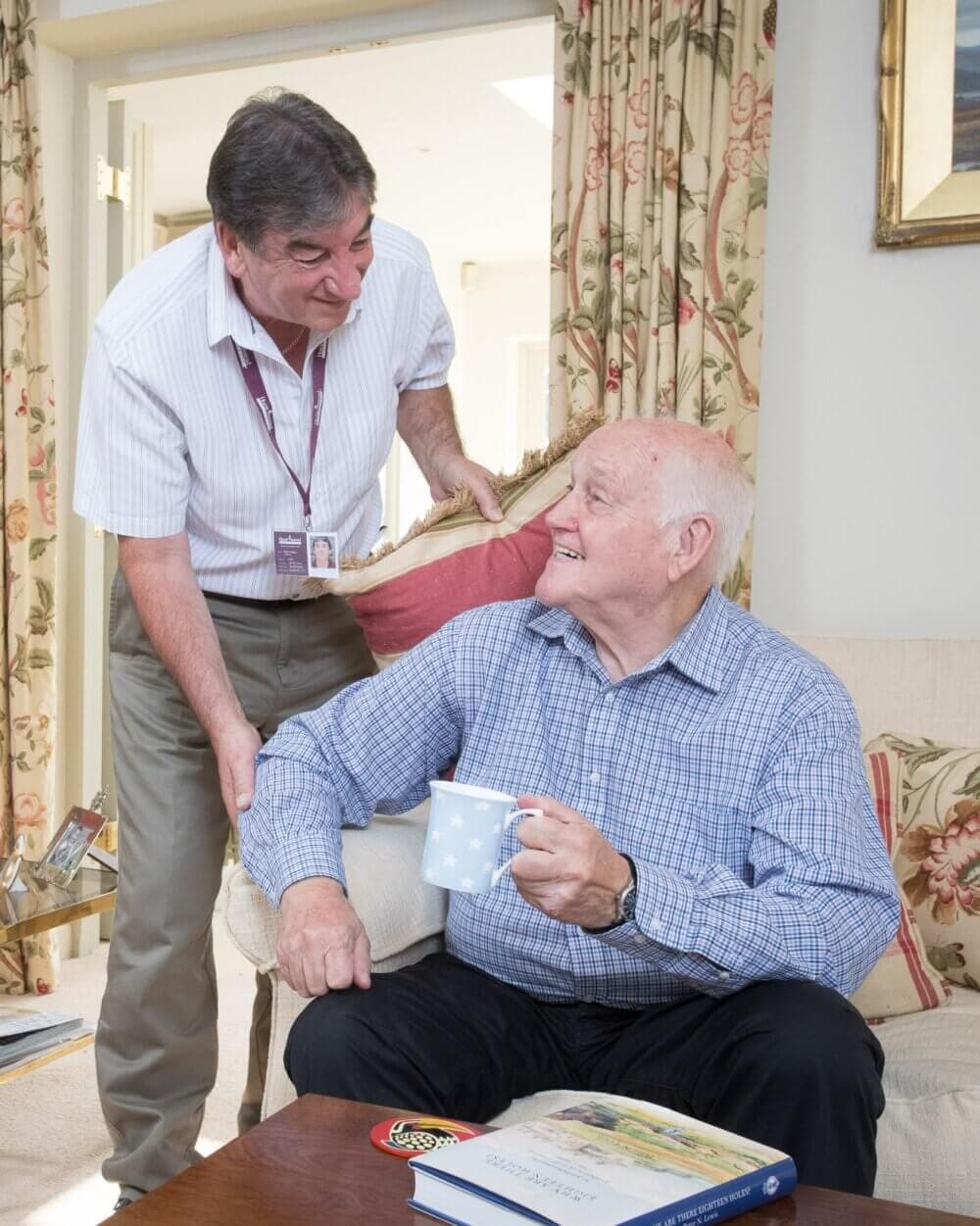 An elderly man holding a mug smiles at a standing man. Both are at home, engaging in conversation. - Home Instead
