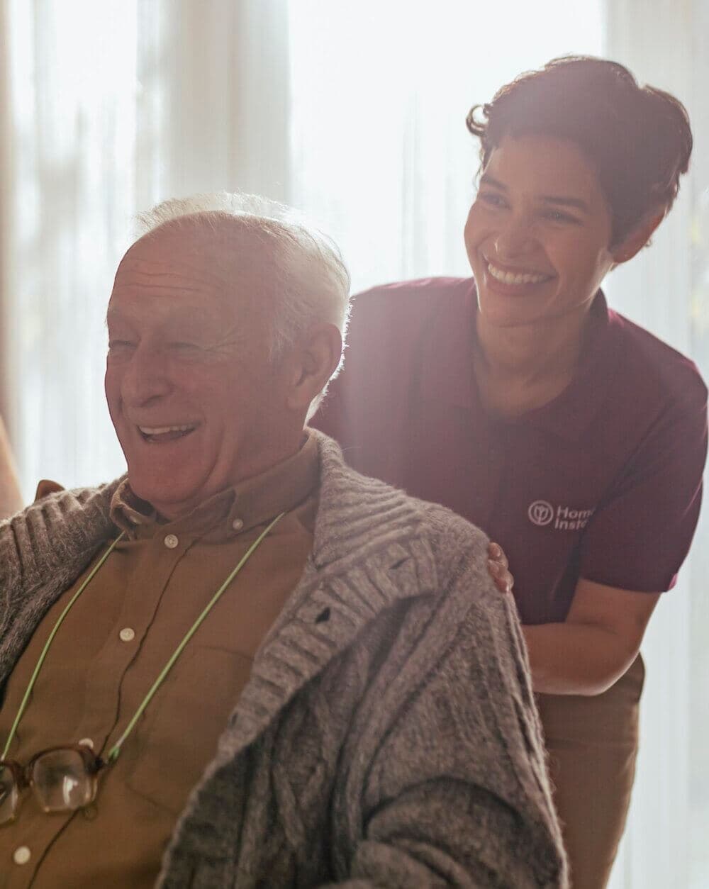 An elderly man laughs with a caregiver standing behind him, smiling, in a warmly lit room. - Home Instead