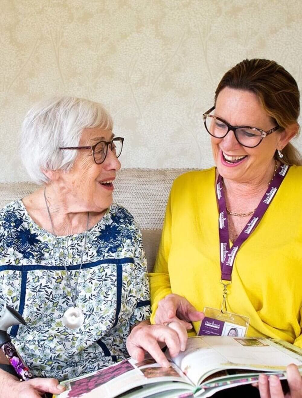 An elderly woman and a caregiver sit together, smiling and looking at a book. - Home Instead