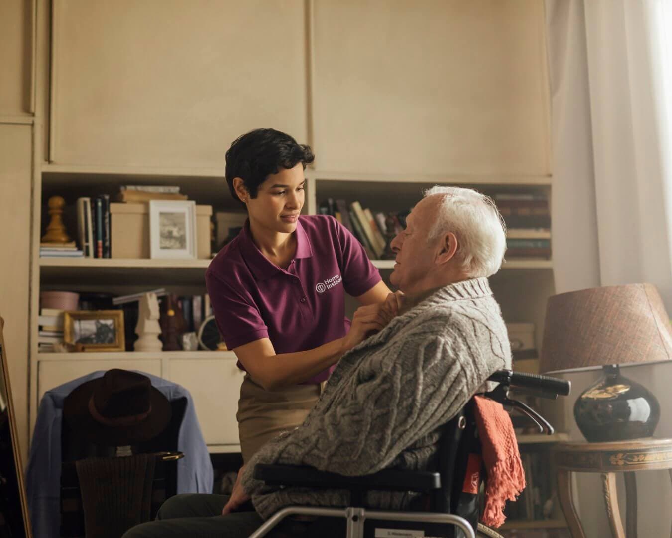 A caregiver in a maroon shirt assists an elderly man in a wheelchair in a cozy, book-filled room. - Home Instead