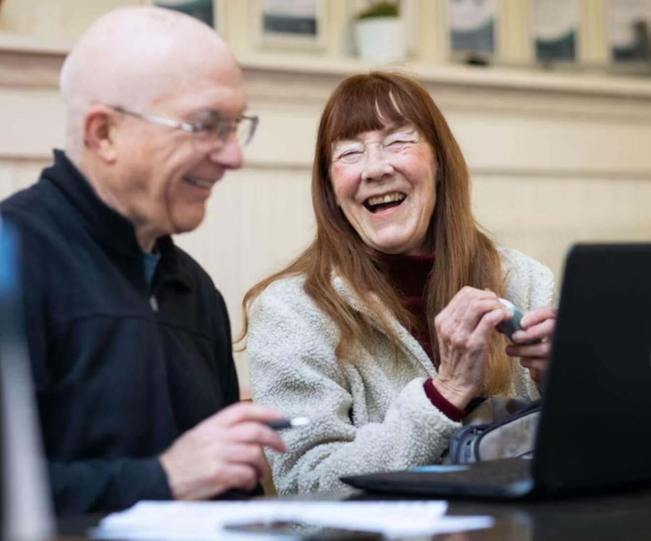 Two older adults sit laughing while working on a laptop at a table. They both appear joyful and engaged. - Home Instead