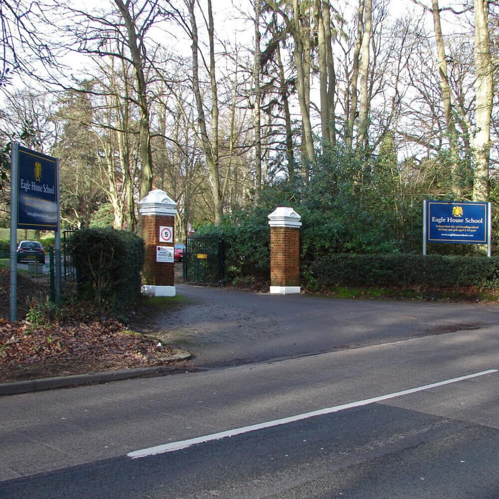 Entrance to Eagle House School with brick pillars, signs, and surrounding trees. Road and autumn leaves in foreground. - Home Instead